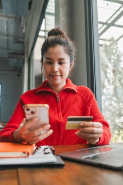 Kırmızı kazaklı bir kadın akıllı telefon ve kredi kartı kullanarak doğal ışık alan bir kafede online alışveriş yapıyor..