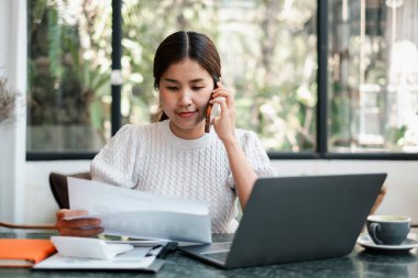 Modern bir ofis ortamında odaklanmış bir kadın, dizüstü bilgisayarla çalışıyor ve telefonla konuşuyor, belgelerle çevrili..