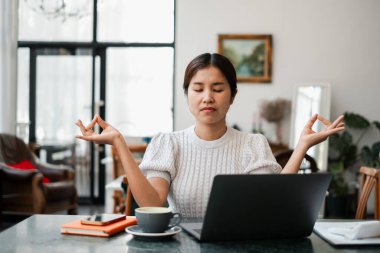 Evde dizüstü bilgisayarla meditasyon yapan bir kadın, rahat bir oturma odasında farkındalık ve iş hayatı dengesini teşvik ediyor..