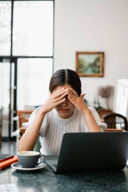 Bir kadın elinde laptop ve kahveyle masada oturuyor, modern bir ofis ortamında stresli görünüyor..