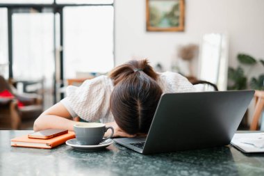 Masasında kafası olan yorgun bir kadın. Etrafı dizüstü bilgisayar, kahve ve defterle çevrili..