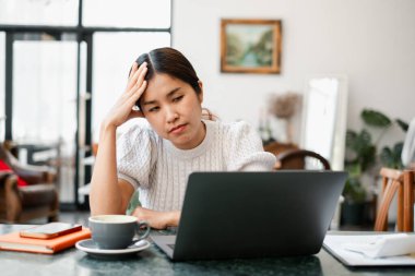 Genç bir kadın bir kafe masasında oturuyor, laptopuyla yakın bir yerde bir fincan kahve ve akıllı telefonla çalışıyor, düşünceli ve odaklanmış görünüyor..