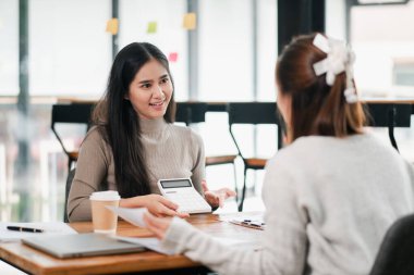 Modern ofis ortamında iş stratejilerini tartışan iki kadın, hesap makinesi ve belge kullanıyorlar..