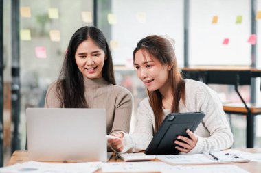 Modern bir ofiste birlikte çalışan iki genç kadın, işbirliği ve beyin fırtınası için bir dizüstü bilgisayar ve tablet kullanıyorlar..