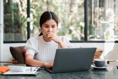 Dışarıda doğal ışık ve yeşillik olan parlak bir kafede dizüstü bilgisayara odaklanan bir kadın..