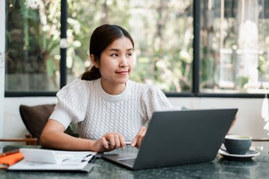 Modern bir kafe ortamında dizüstü bilgisayarına odaklanan genç bir kadın, doğal ışık ve yeşillik ile çevrili, yanında bir fincan kahveyle.