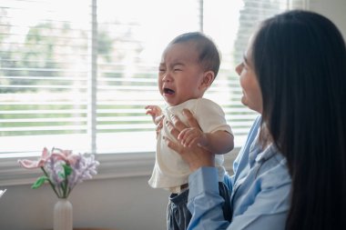 Bir anne ağlayan bebeğini pencere perdeleri ve bir vazoyla parlak bir odada tutar, hassas bir anı yakalar..
