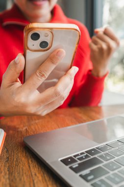 Kırmızı kazaklı bir kadının laptopun yanında akıllı telefon kullanırken, rahat bir kafede modern teknoloji kullanımının görüntülerini çekerken..