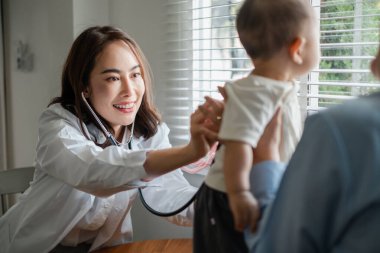 Güneşli bir klinikte bebeğin sağlığını kontrol etmek için steteskop kullanan gülümseyen bir çocuk doktoru..