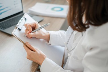 Modern ofis ortamında not alan kadın bir doktor. Bilgisayarı ve sağlık raporları görünür..