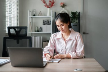 Masasında laptopuyla parlak bir ofis ortamında notlar yazan odaklanmış genç bir kadın..