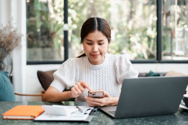 Akıllı telefon ve dizüstü bilgisayarla modern ofis ortamında birden fazla iş yapan genç bir kadın..