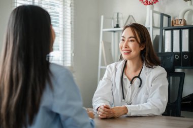 Beyaz önlüklü bir kadın doktor, parlak ve modern bir ofiste hastasına danışırken gülümsüyor..