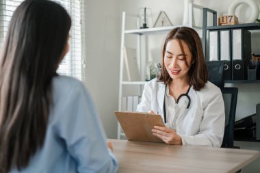 Beyaz önlüklü kadın doktor modern ofis ortamında bir hastaya danışarak tıbbi kayıtları tartışıyor..