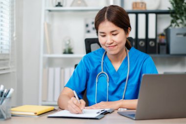Mavi önlüklü bir hemşire elinde dizüstü bilgisayarla bir masanın üzerine yazı yazıyor ve profesyonel bir sağlık hizmeti ortamı sergiliyor..