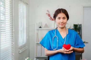 Mavi önlüklü bir hemşire kalp şeklinde bir nesne tutuyor. Parlak bir sağlık ofisinde şefkat ve ilgiyi simgeliyor..