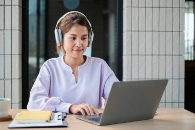 Kulaklık takan genç bir kadın, modern bir ofiste dizüstü bilgisayarla çalışıyor. Uzak çalışma, teknoloji ve üretkenlik temaları için ideal.