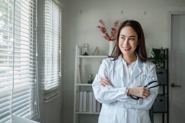 Beyaz önlüklü gülümseyen kadın doktor, elinde stetoskop, parlak bir muayenehanede güvenle duruyor..