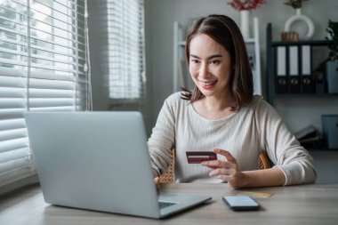 Neşeli bir kadın bilgisayarını ve kredi kartını iyi aydınlatılmış bir ofis ortamında online alışveriş yapmak için kullanır..