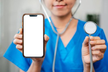 Mavi önlüklü bir sağlık çalışanı elinde boş bir ekran ve stetoskop olan bir akıllı telefon tutuyor. Tıbbi teknolojiyi simgeliyor..