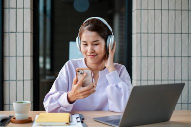 Kulaklık takan, bilgisayarlı ve kahveli bir masada akıllı telefon kullanan, modern bir çalışma alanında müziği seven bir kadın..