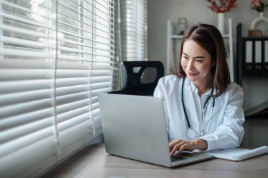 Beyaz önlüklü kadın doktor masadaki parlak ofiste dizüstü bilgisayarını kullanarak modern sağlık ve teknolojiyi sergiliyor..