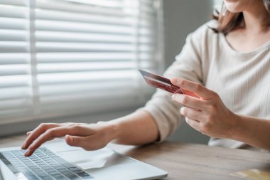 Bir kadın bilgisayarından alışveriş yapmak için kredi kartı kullanıyor. Ev ortamında panjurları olan bir pencerenin yanında oturuyor..