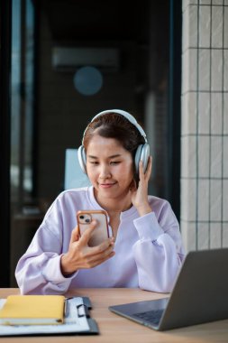 Kulaklık takan, dizüstü bilgisayarı ve not defteri olan bir masada akıllı telefon kullanan, modern bir ofis ortamında müzikten hoşlanan gülümseyen bir kadın..