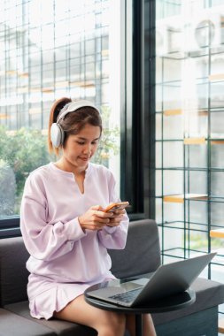 Kulaklık takan genç bir kadın, modern bir kafede akıllı telefon kullanıyor ve masada dizüstü bilgisayarı var..