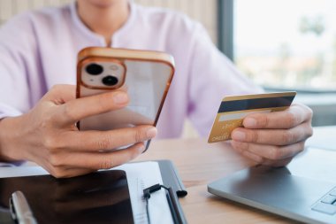 Elinde akıllı telefon ve kredi kartı olan biri ev ofisinin masasında dizüstü bilgisayarla online alışveriş yapıyor..