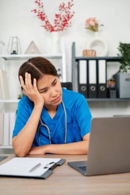 Mavi önlüklü, ofis malzemeleri ve dekorasyonla çevrili bir laptopla bir masada oturan bitkin bir sağlık görevlisi..