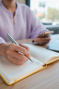 Akıllı bir telefon tutarken deftere yazan bir kadının, parlak bir ofis alanı içerisindeki yakın çekimi..