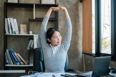 Çizgili gömlekli genç bir kadın çalışma masasına uzanır. Etrafı kitap ve belgelerle çevrilidir. Modern bir çalışma alanında..