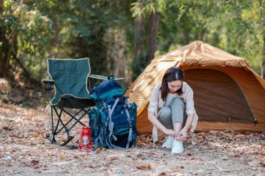 Orman ortamında bir çadırın yanında ayakkabılarını bağlayan bir kadın. Etrafı kamp malzemeleri ve doğa ile çevrili..