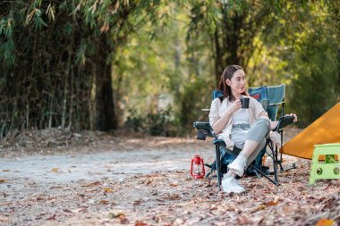 Bir kadın, sonbahar yaprakları ve ağaçlarla çevrili bir çadırın yanında kamp koltuğunda oturup kahvenin tadını çıkarıyor..