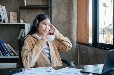 Modern ofis ortamında kahve ve müziğin keyfini çıkaran günlük kıyafetli genç bir kadın. Etrafı belgeler ve dizüstü bilgisayarla çevrili..