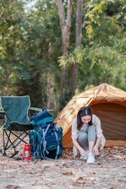 Kamp malzemeleri ve doğa ile çevrili bir orman kampında çadırın yanında ayakkabılarını bağlayan bir kadın..