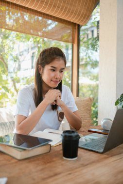 Genç bir kadın dizüstü bilgisayarıyla çalışıyor. Parlak, rahat bir ev ofisinde. Kahvesi var, etrafı yeşilliklerle çevrili..