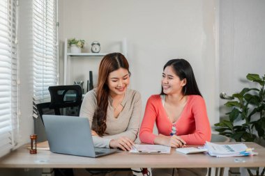 İki kadın ofis masasında birlikte çalışıyor, gülümsüyor ve modern bir ortamda dizüstü bilgisayarla belgeleri tartışıyorlar..