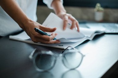 Gözlüklü bir masadaki belgelere yazı yazan ve profesyonel bir ofis ortamı sergileyen ellerin yakın çekimi.