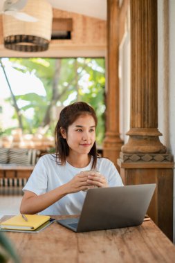 Beyaz tişörtlü genç bir kadın, yeşilliklerle çevrili, güneşli bir kafede, ahşap bir masada dizüstü bilgisayar kullanıyor..