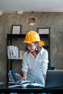 Sarı şapkalı kadın mimar modern bir ofisin planlarını inceliyor, profesyonel çalışma ortamını gözler önüne seriyor..