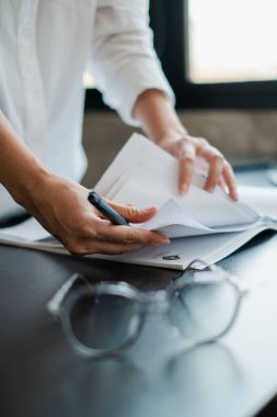 Modern bir ofis ortamında, gözlüklü ve kalemli kağıtları düzenleyen ellerin yakın çekimi..