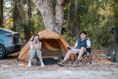 Bir çift, doğa tarafından çevrelenmiş, çadırlarının ve arabalarının yanında, ormanda huzurlu bir kamp yolculuğunun tadını çıkarıyor..