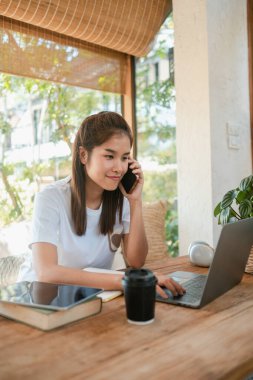 Genç bir kadın, dizüstü bilgisayarı üzerinde çalışarak ve telefonuyla konuşarak rahat, aydınlık bir ofis ortamında birden fazla iş yapar..