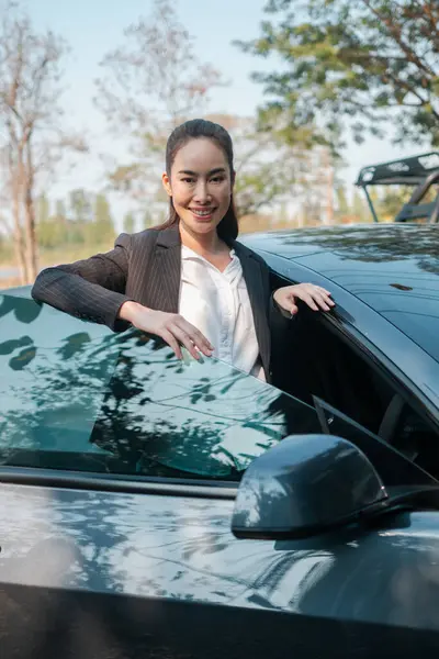 İş kıyafeti giymiş profesyonel bir kadın, etrafı doğayla çevrili bir arabanın yanında kendinden emin bir şekilde duruyor..