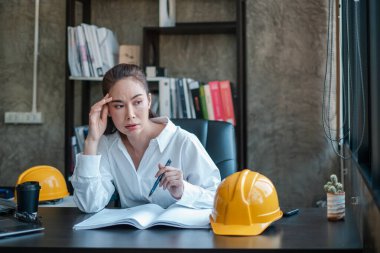 Kadın mimar masasında güvenlik kaskları, kitaplar ve modern ofis dekorlarıyla planları analiz ediyor..