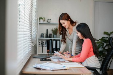 İki kadın bir ofis masasında birlikte çalışıyor, belgeleri inceliyor ve modern, parlak bir çalışma alanında dizüstü bilgisayar kullanıyor..