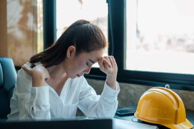 Beyaz gömlekli kadın bir mühendis masasında stresli görünüyor, yanında sarı bir kaskla, modern ofis ortamında..
