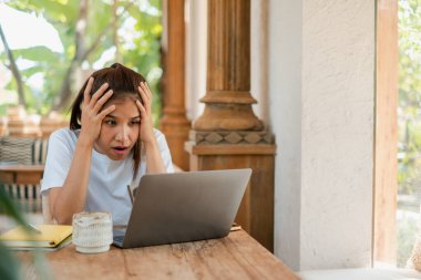 Genç bir kadın evde dizüstü bilgisayarıyla çalışırken stresli görünüyor. Etrafı kahve ve ahşap bir masada defter ile çevrili..
